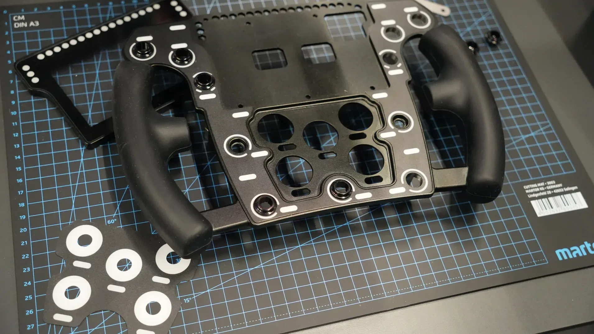 Un volant Pokornyi Engineering en kit, ses pièces étalées sur un tapis de découpe du stand. De quoi monter son propre volant.